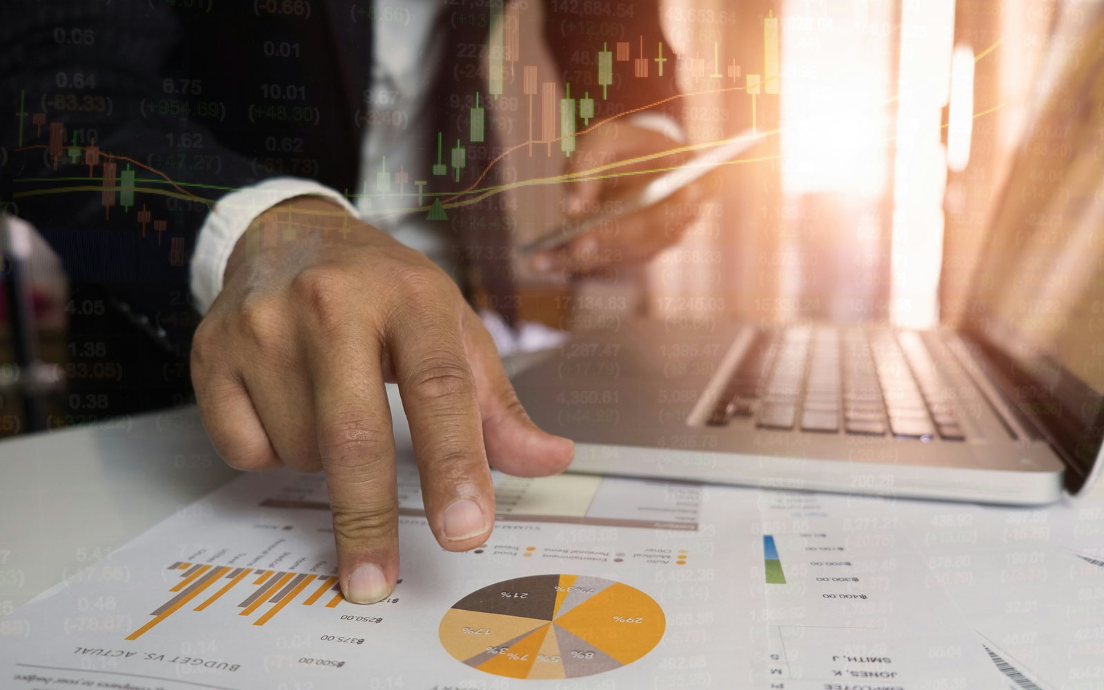A hand pointing at printed financial charts beside a laptop, with market data overlaid