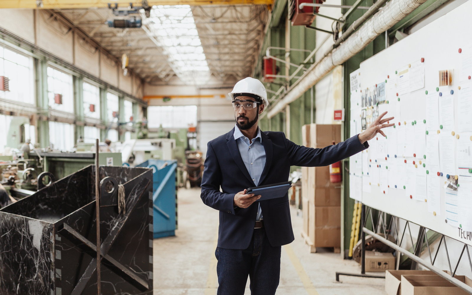 A plant manager in a hard hat reviewing a production board on a factory floor